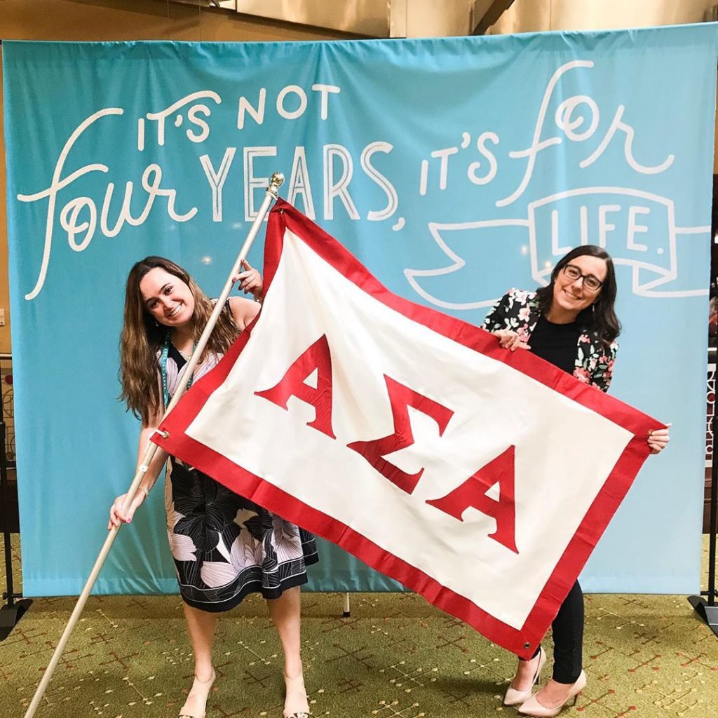 Two women holding a flag with Alpha Sigma Alpha's letters on it. They are standing in front of a banner that reads, "it's not for four years, it's for life."